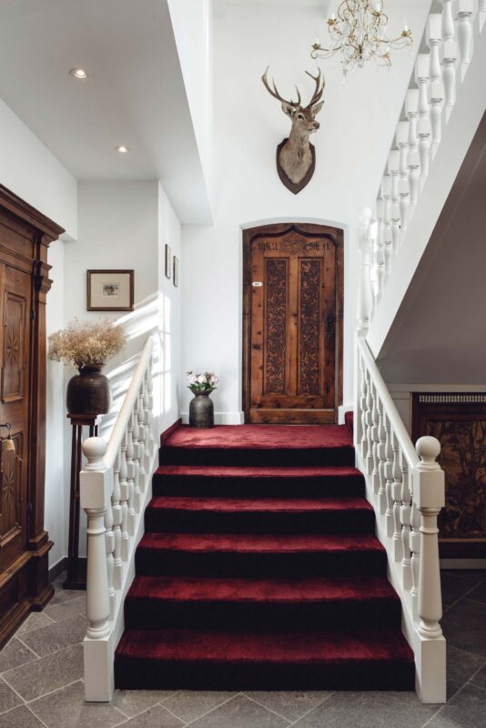 Elegant staircase with red carpet leading to a wooden door, adorned with a deer head mount and decorative furnishings.
