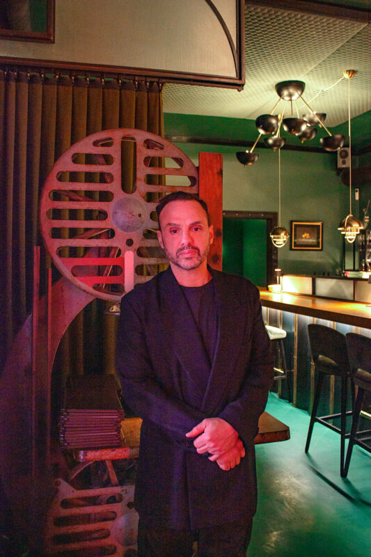 Man in a black suit stands in a dimly lit bar with vintage film reel decor and green walls.