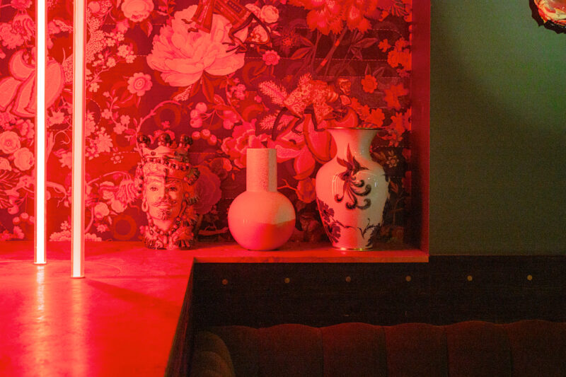 Colorful vases and a decorative bust on a shelf against a floral-patterned wall, illuminated with red light.