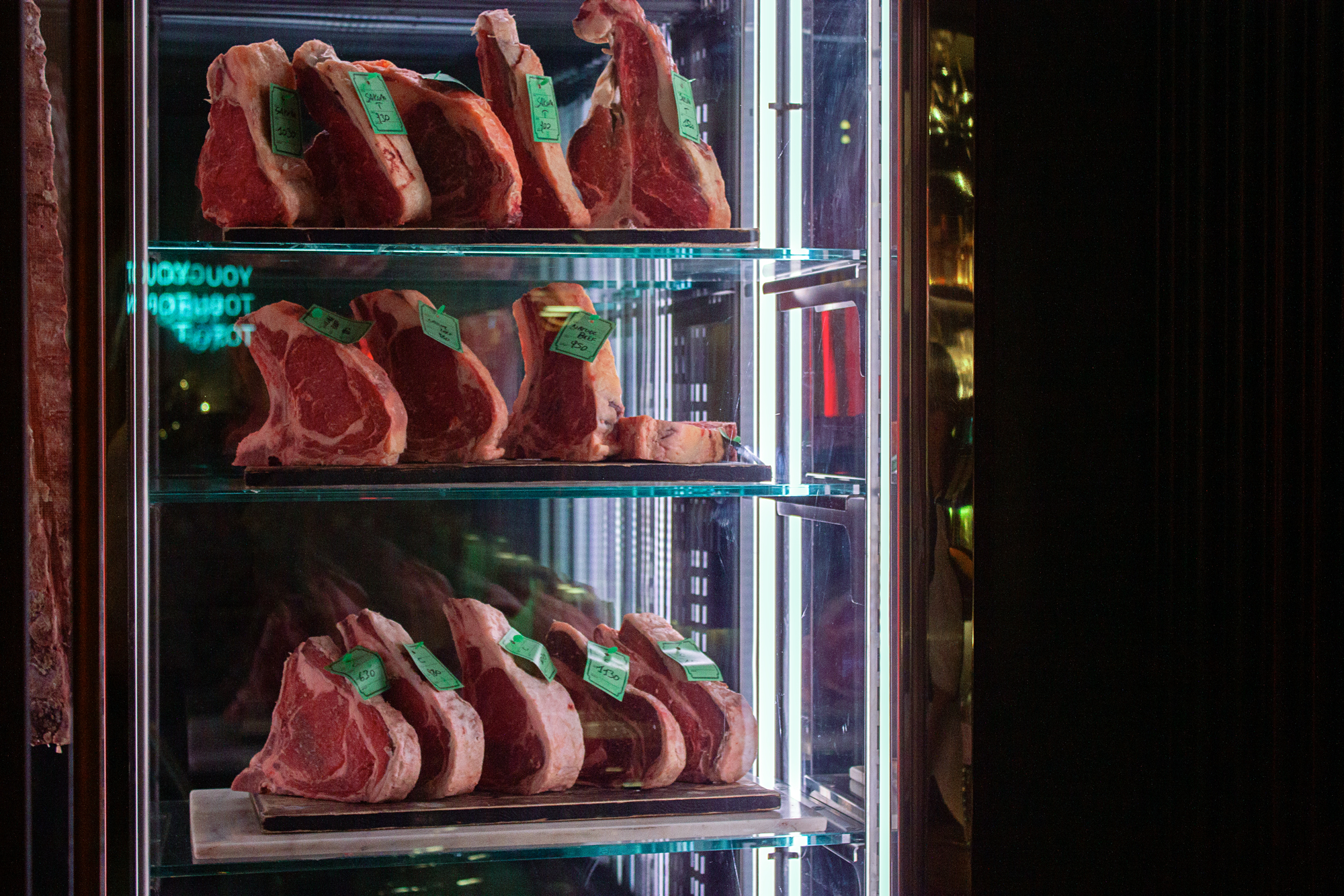 Display case with several cuts of meat, each labeled with green tags, illuminated in a restaurant setting.
