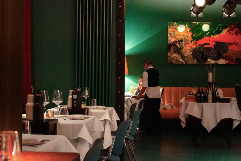 Elegant restaurant interior with green walls, featuring tables set with white tablecloths, a waiter attending to a guest, ...
