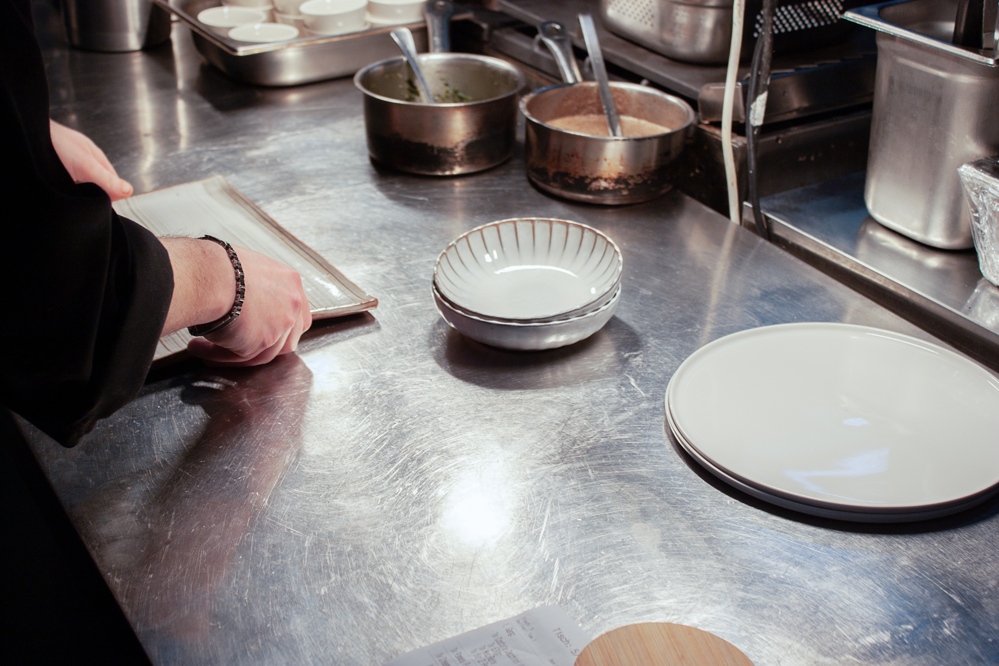 A chef's hands prepare a menu on a stainless steel countertop with empty plates and bowls in the background.