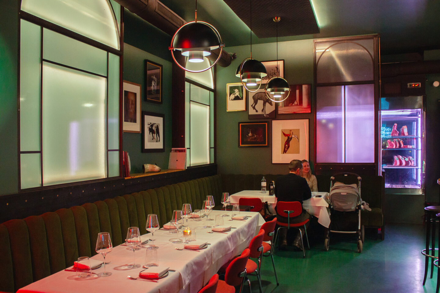 Elegant restaurant interior with green walls, red chairs, and dim lighting; a couple dining with a stroller nearby.