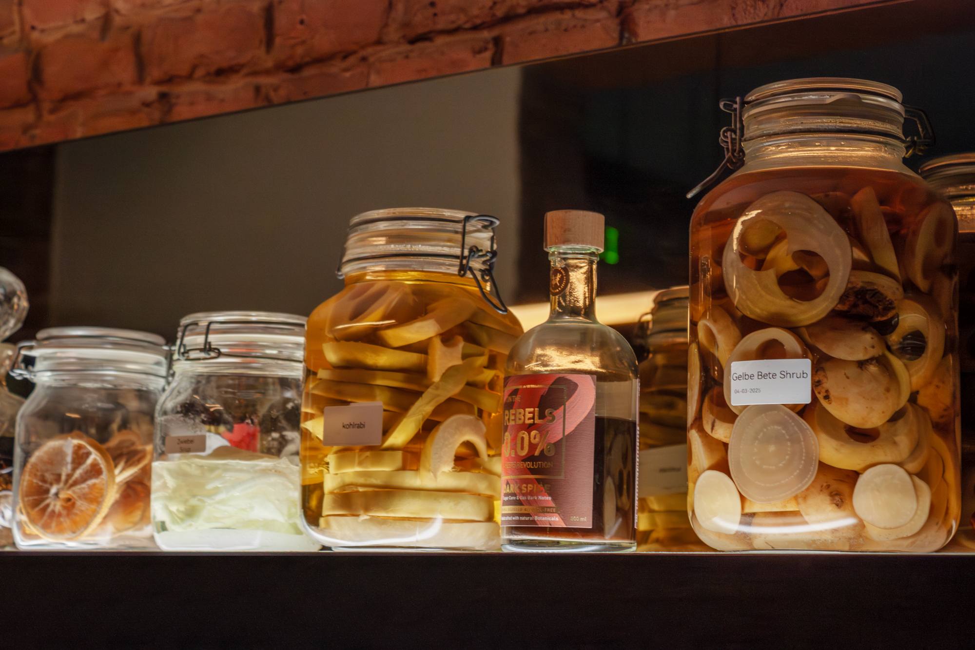 Glass jars filled with various pickled vegetables and fruits, including sliced onions and citrus, displayed on a shelf.