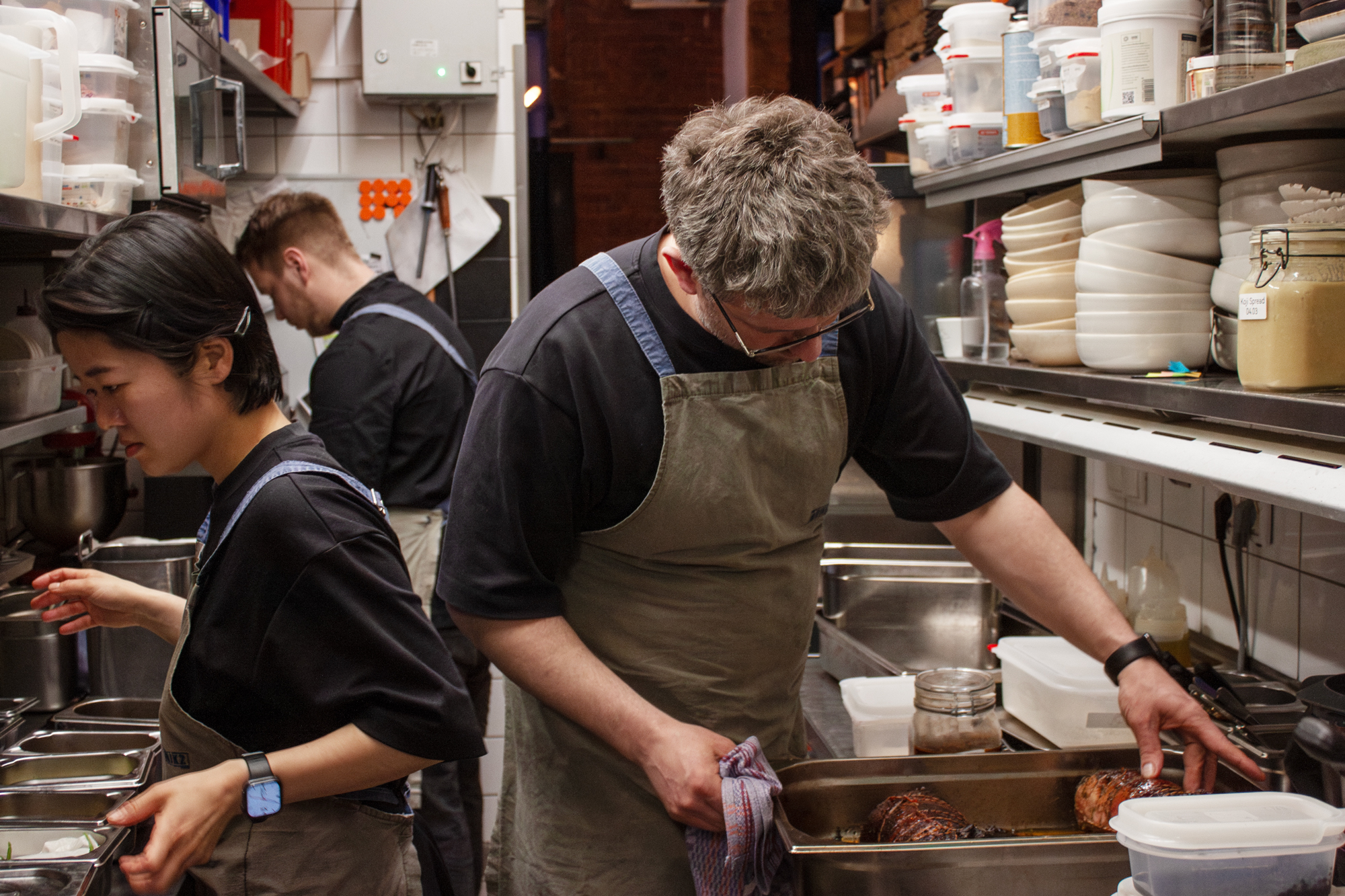 Two chefs in an industrial kitchen, one inspecting a tray of food while the other prepares ingredients.