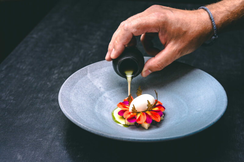 A hand pours a creamy sauce over a dessert featuring flowers and a scoop of ice cream on a gray plate.