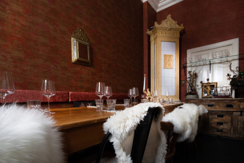 Elegant dining setup with a wooden table, glassware, and fur throws, against a richly textured red wall and ornate cabinet.