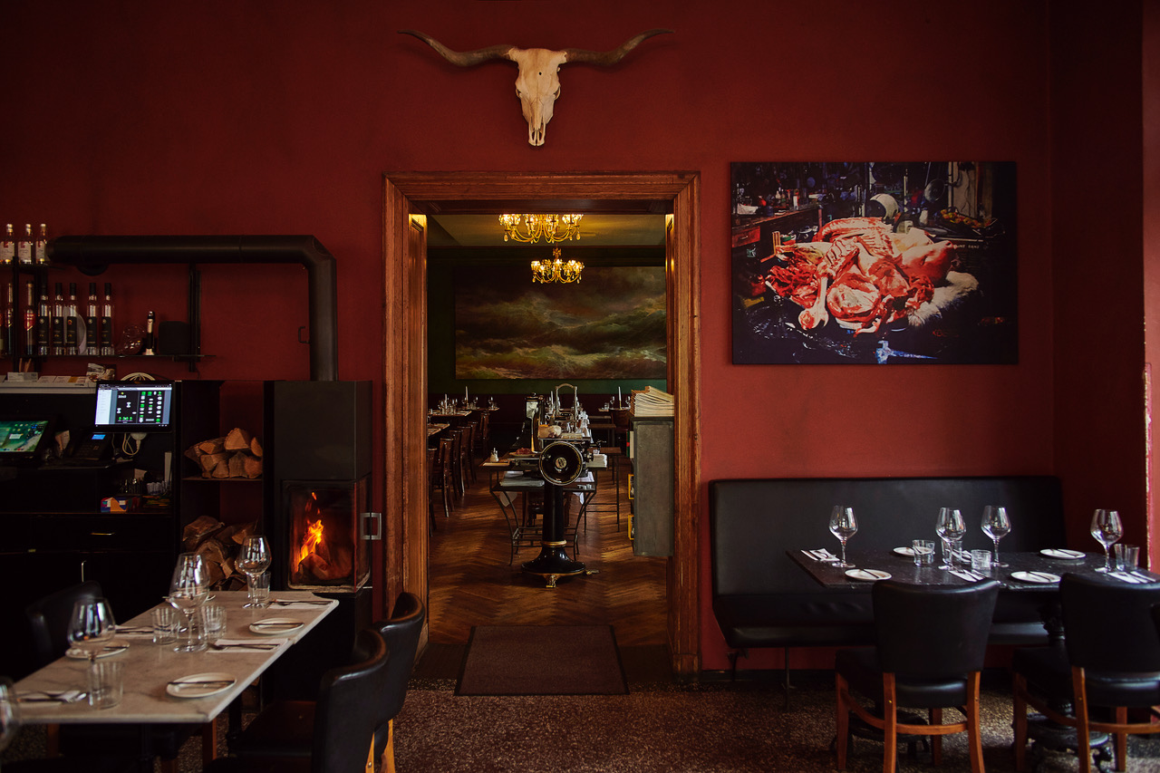 Interior of a restaurant with red walls, a bull skull, and a meat-themed artwork, featuring tables set for dining.