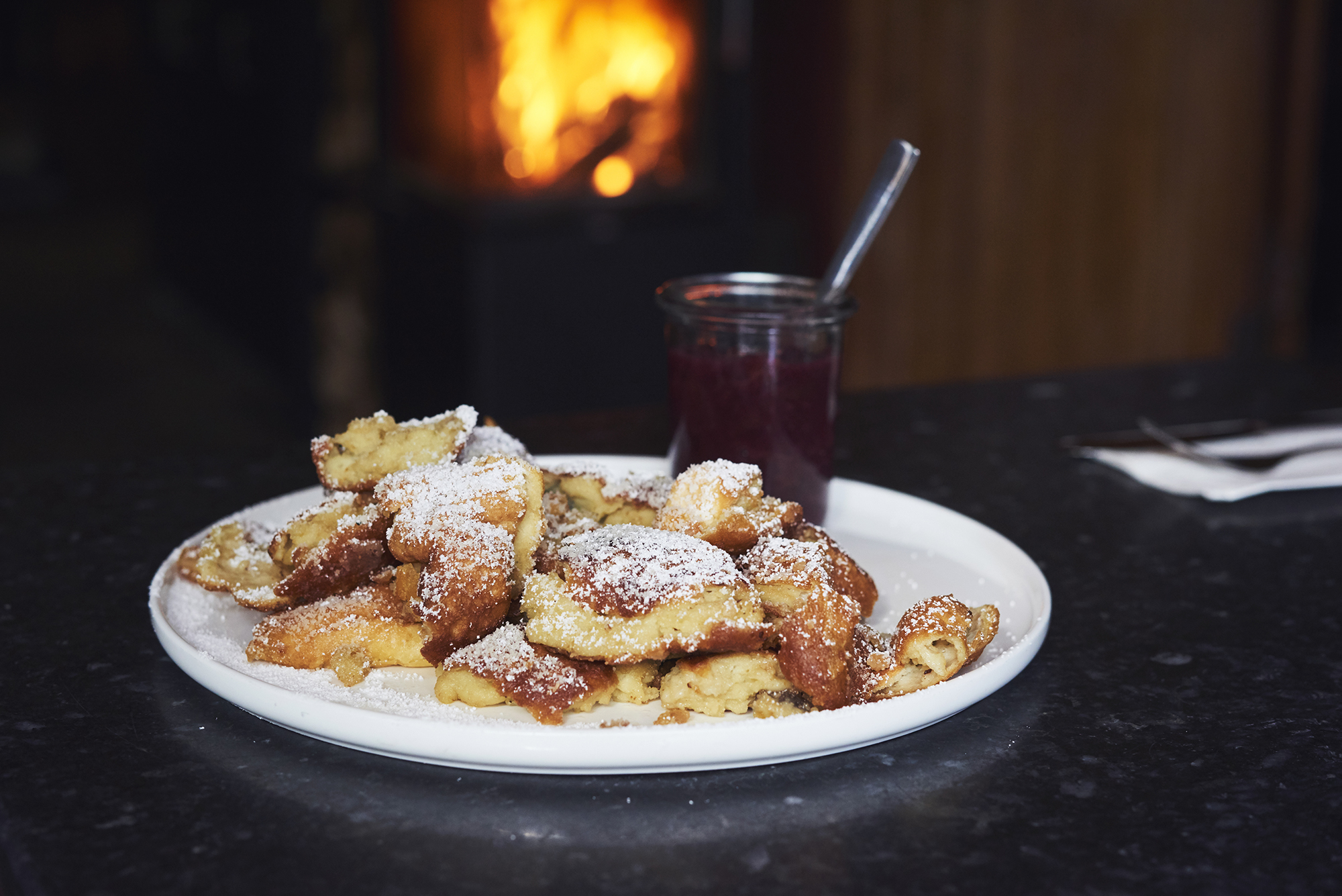 Plate of Kaiserschmarrn dusted with powdered sugar, served with a jar of berry jam, against a warm, glowing fireplace.