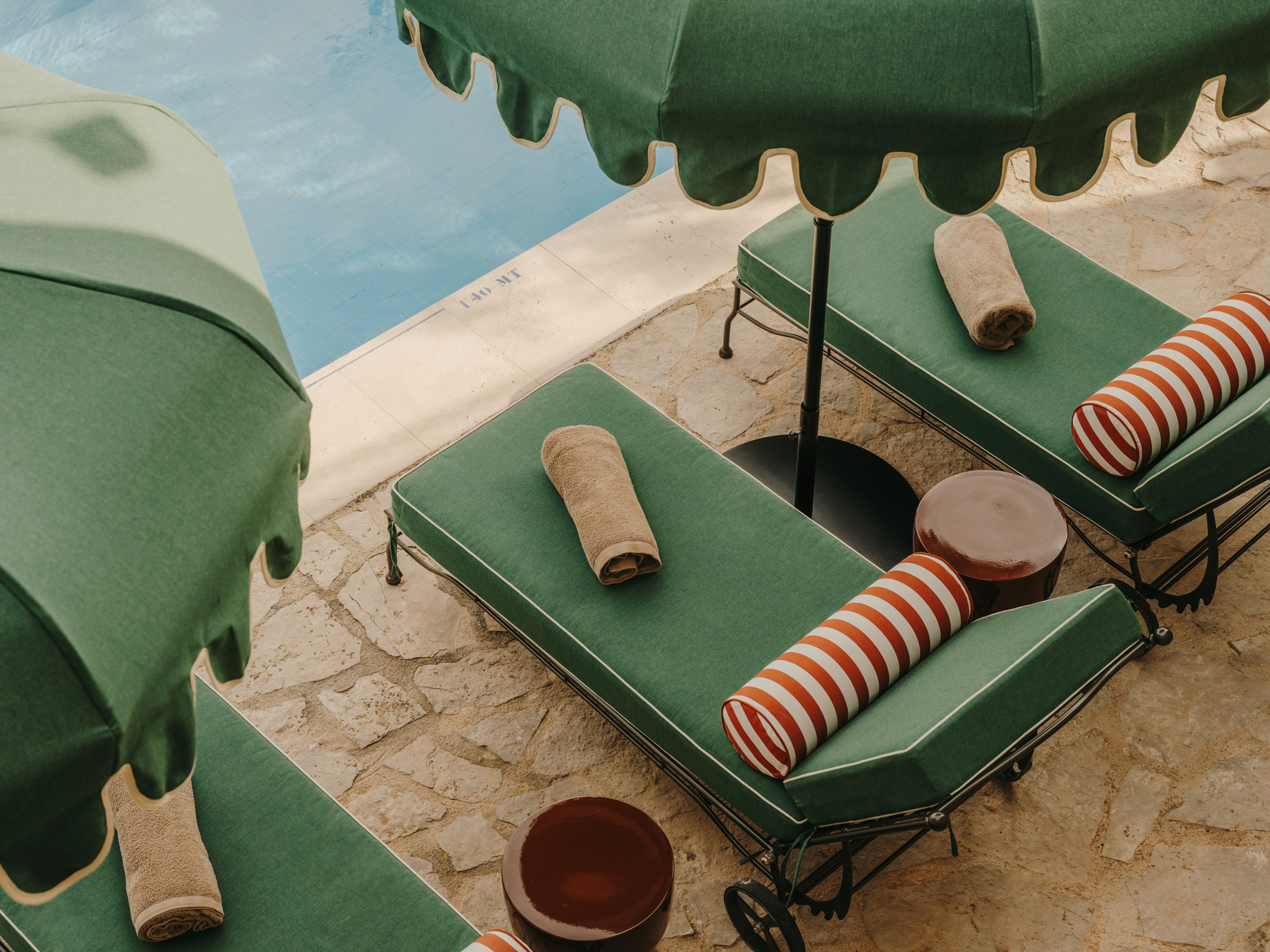 Green lounge chairs with striped cushions and rolled towels under green umbrellas by a poolside.
