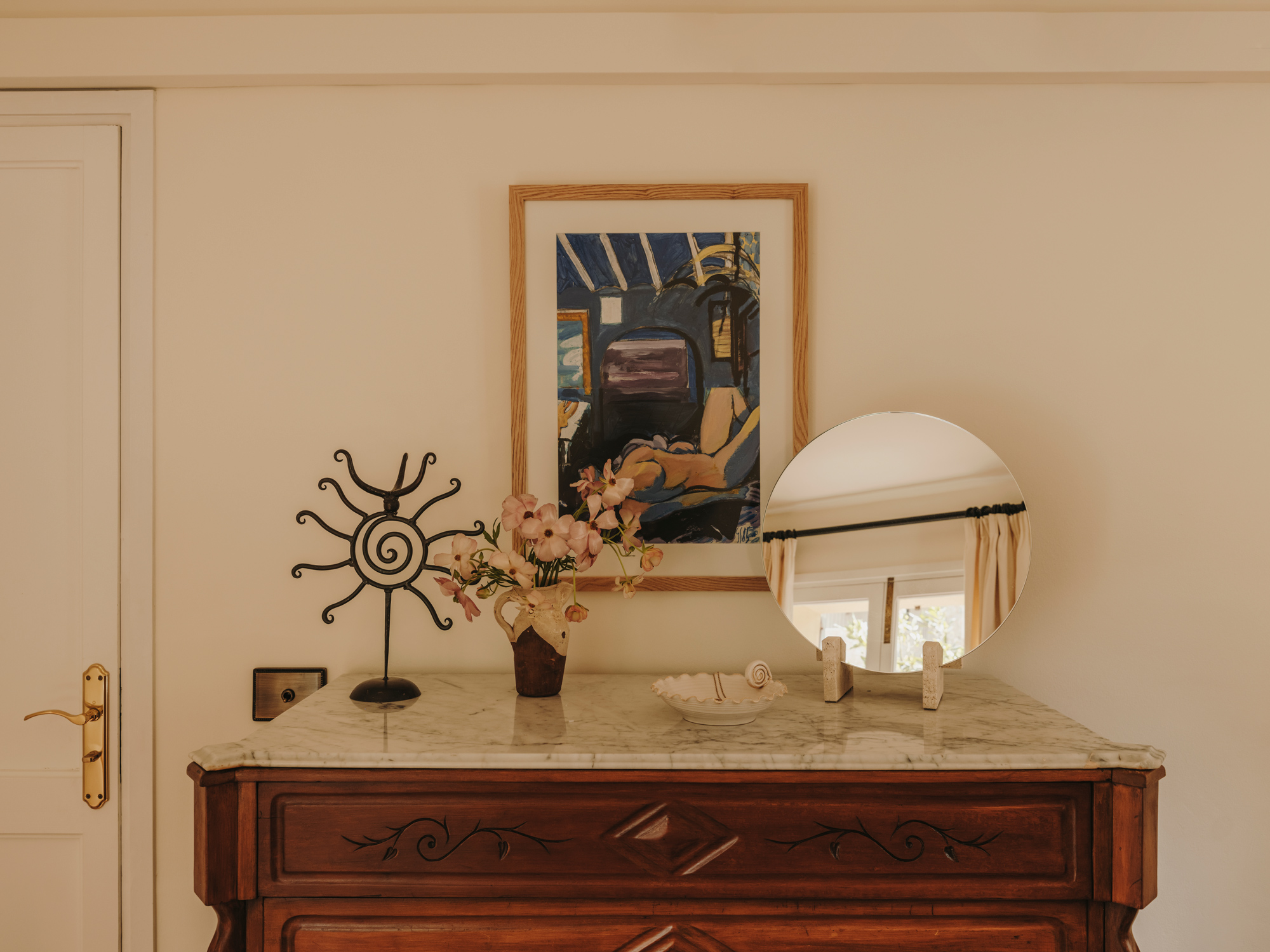 Wooden dresser with marble top, featuring a framed painting, a vase of flowers, a decorative mirror, and a metal sculpture.