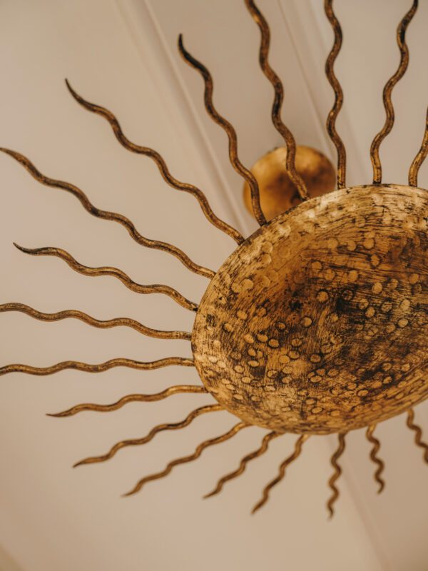 Gold sun-shaped chandelier with wavy rays, hanging from a white ceiling.