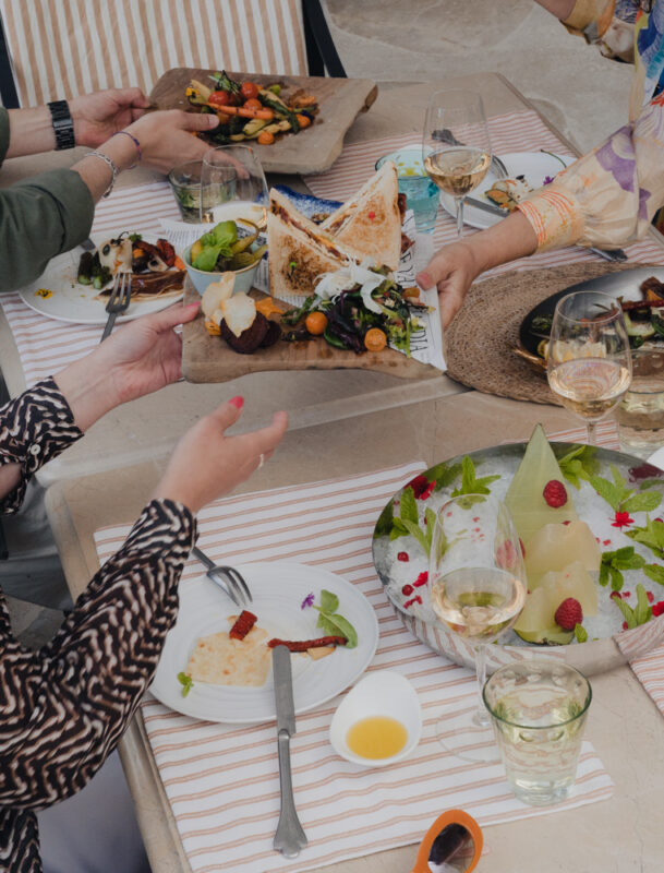 A table set with various gourmet dishes, wine glasses, and hands exchanging plates, showcasing a vibrant dining experience.