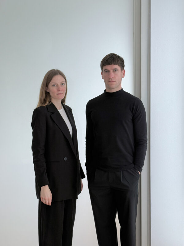 A man and woman stand side by side against a light background, both dressed in black attire.