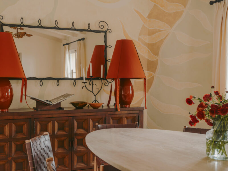 Warm-toned interior featuring a wooden sideboard with a large mirror, red lamps, and a vase of flowers on a round table.