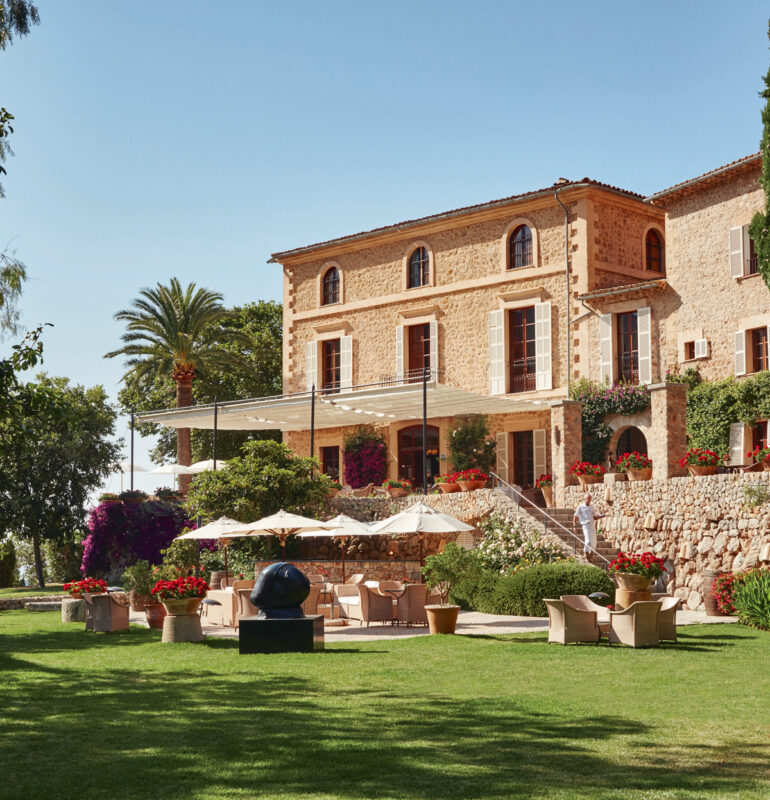 Elegant stone building surrounded by lush greenery and colorful flowers, featuring outdoor seating and umbrellas.