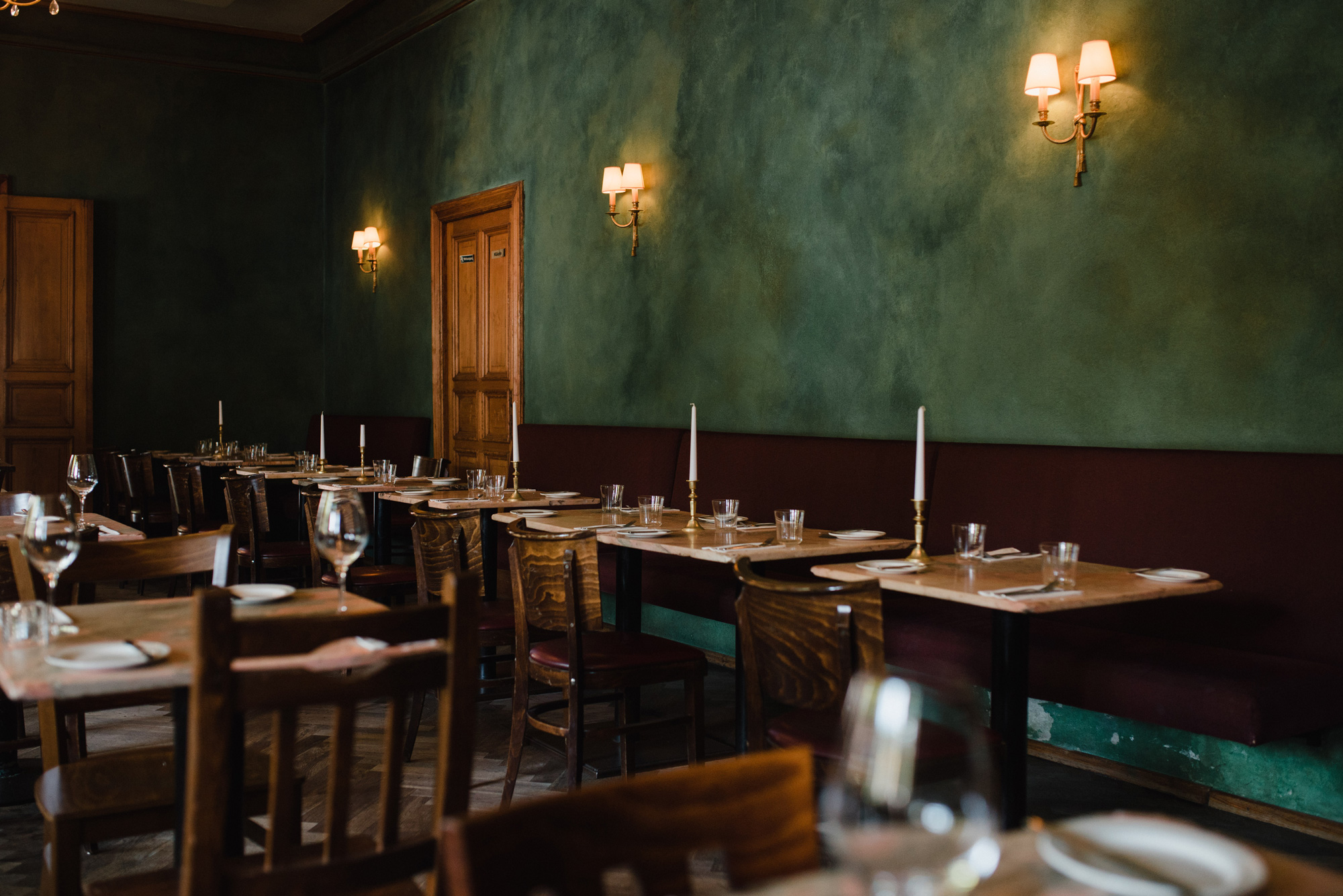 Elegant restaurant interior with green walls, wooden tables set with glassware and candles, and warm lighting.