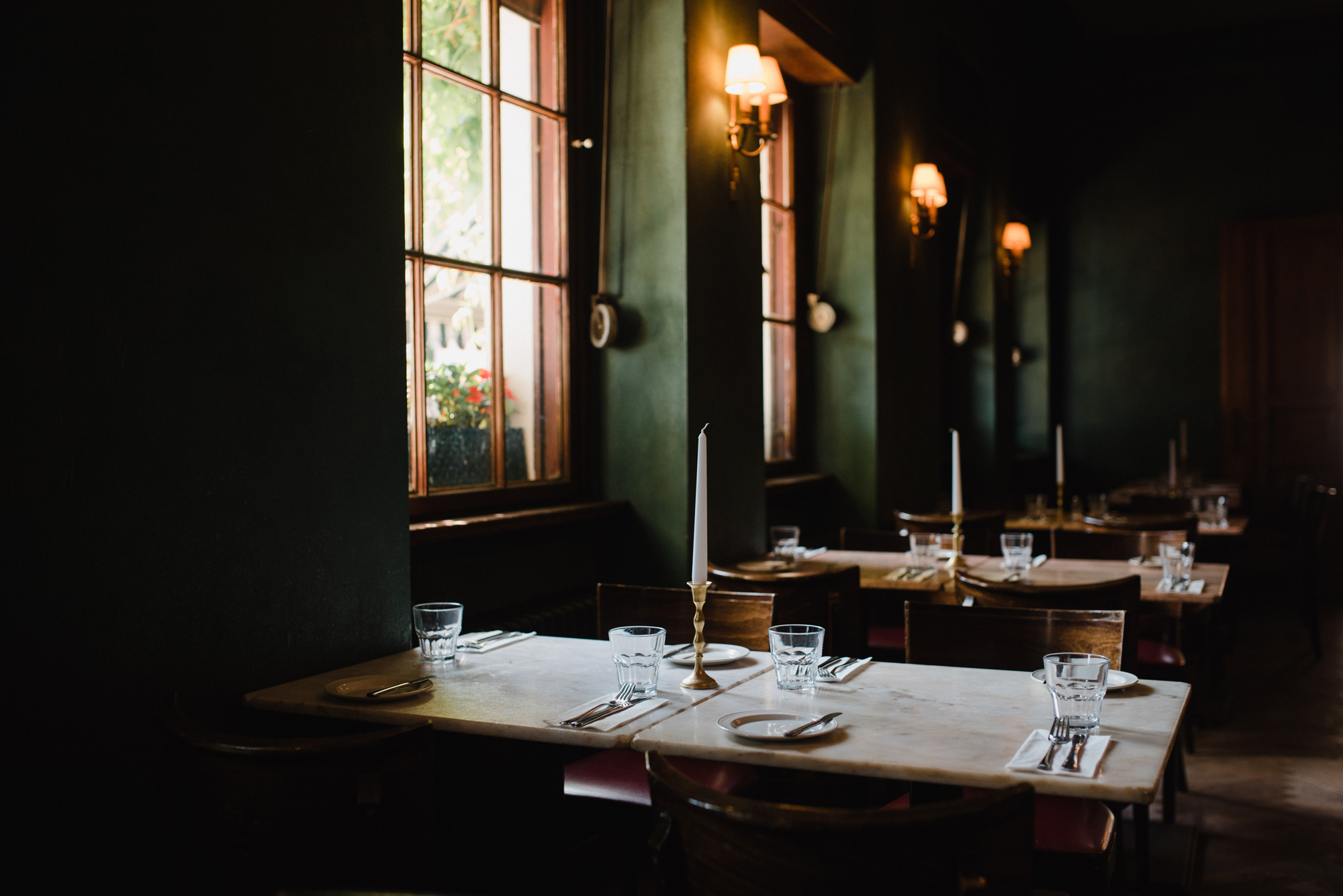 Elegant restaurant interior with dark green walls, wooden tables set with glassware and candles, and large windows.