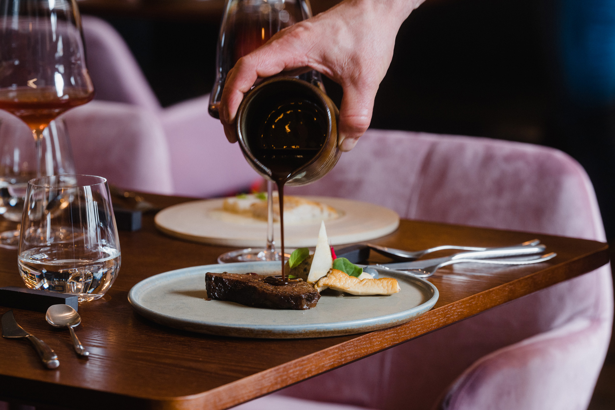 A hand pours sauce over a plate with beef cheek, celery, and mushroom, set on a wooden table in a restaurant.