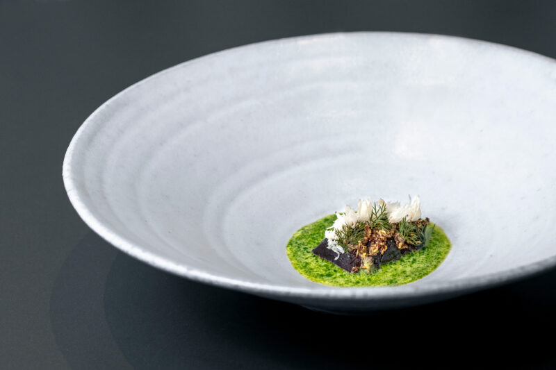 A white bowl containing a vibrant green sauce, topped with pieces of roasted beetroot, herbs, and crunchy elements.