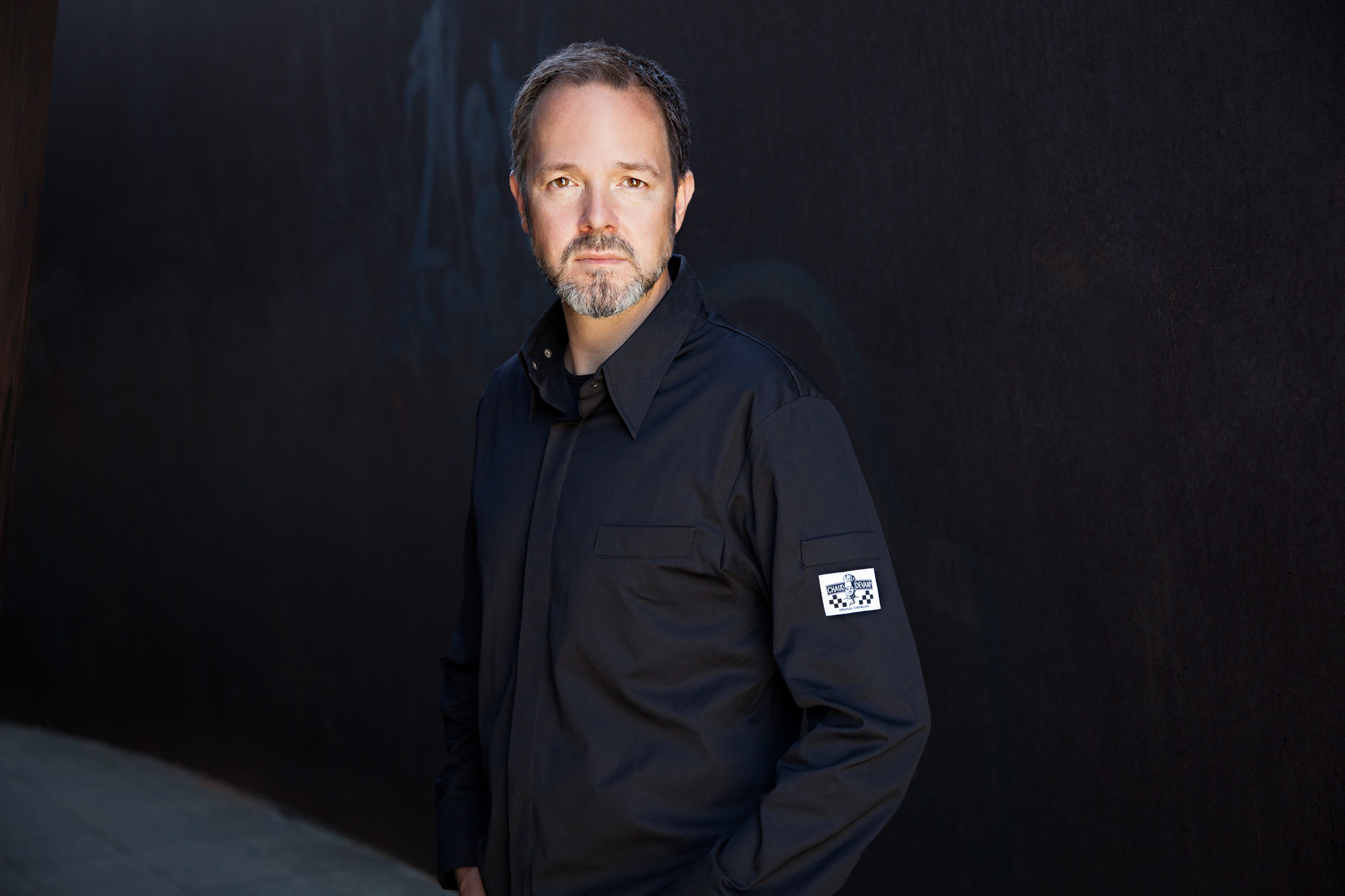 Man with a beard wearing a black shirt stands confidently against a textured dark background.