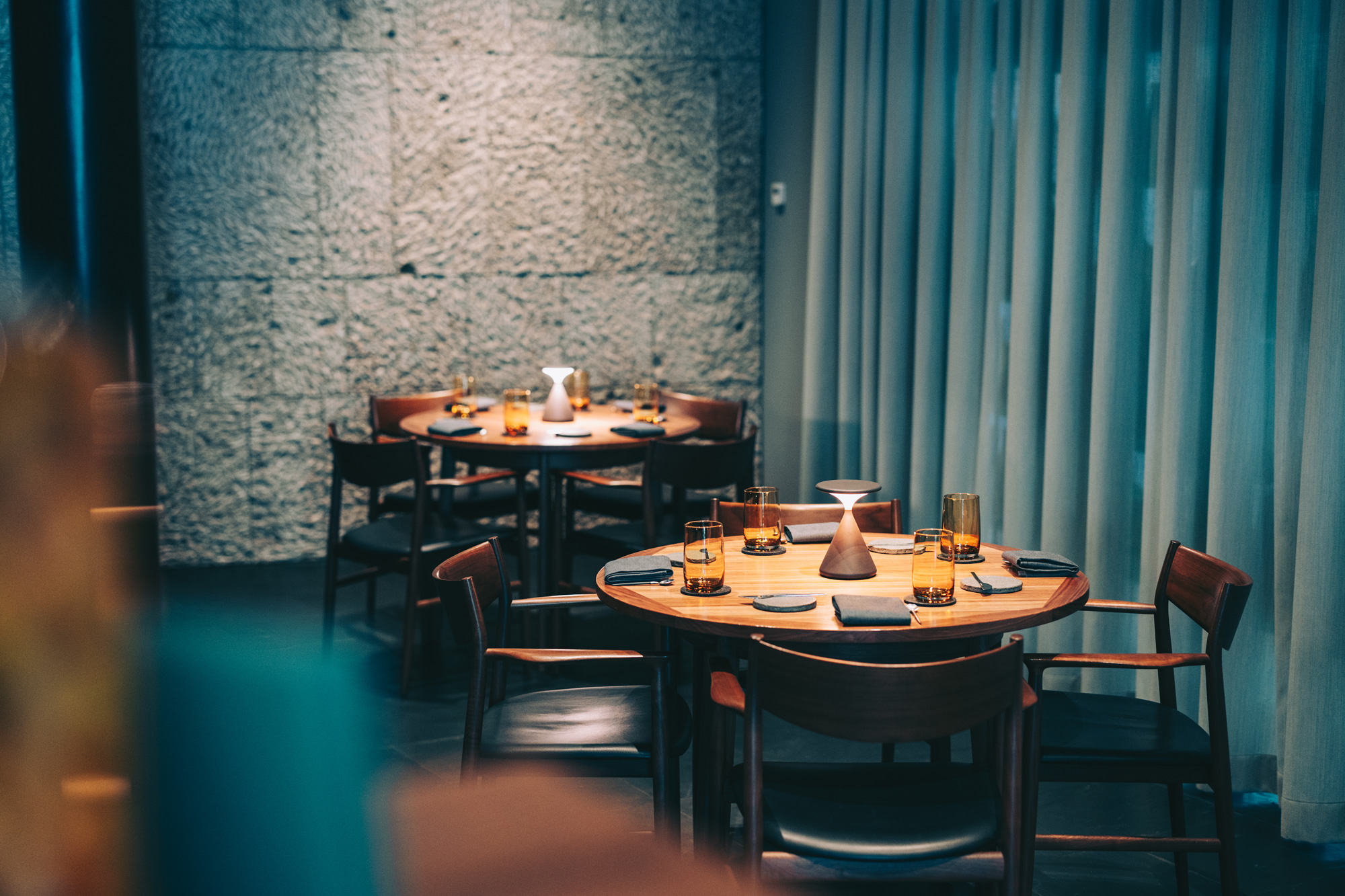 Elegant dining setup with wooden tables, dark chairs, and amber glasses, against a textured wall and soft curtains.