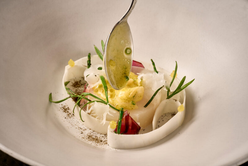 Delicate dish featuring grilled cod with shellfish flavors, garnished with herbs and a drizzle of sauce.