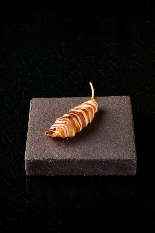 A single, elongated food item drizzled with sauce, placed on a textured dark gray plate.