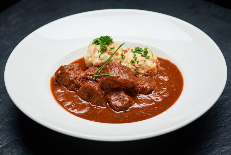 Rich beef goulash in a savory sauce, served with two dumplings and garnished with chives.