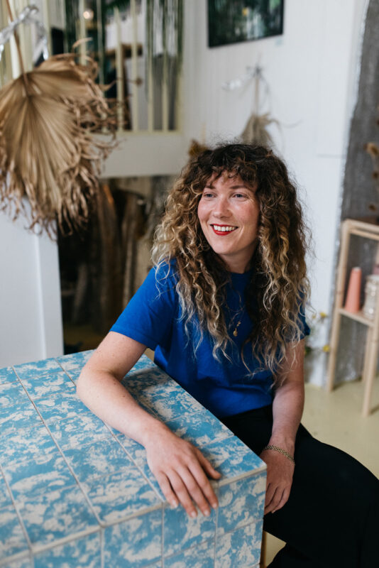 Woman with curly hair in a blue shirt smiles while sitting at a patterned table in a stylish interior.