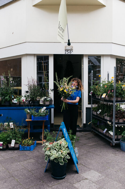 Woman with curly hair holding flowers outside a flower shop, surrounded by plants and a blue display sign.