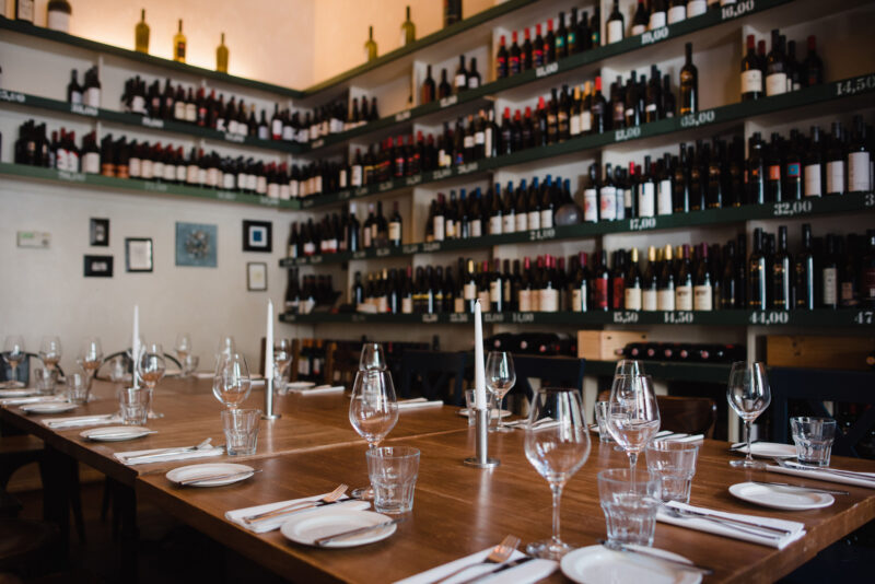 Elegant restaurant interior with a wooden table set for dining, surrounded by shelves filled with wine bottles.