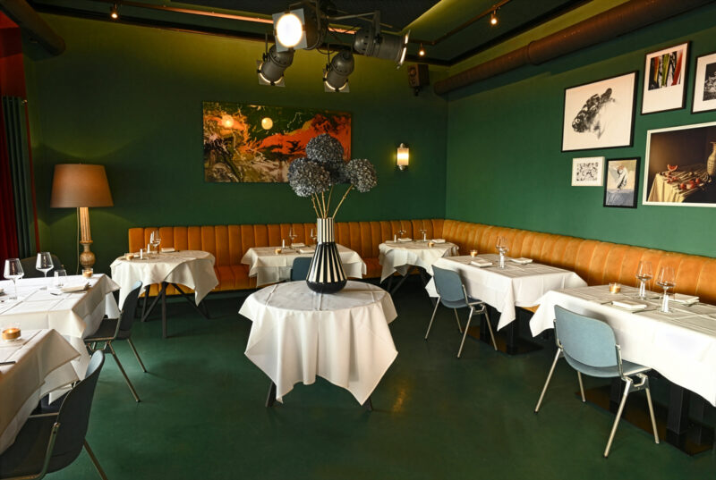 Elegant restaurant interior with green walls, white tablecloths, and a central floral arrangement, featuring artwork on th...