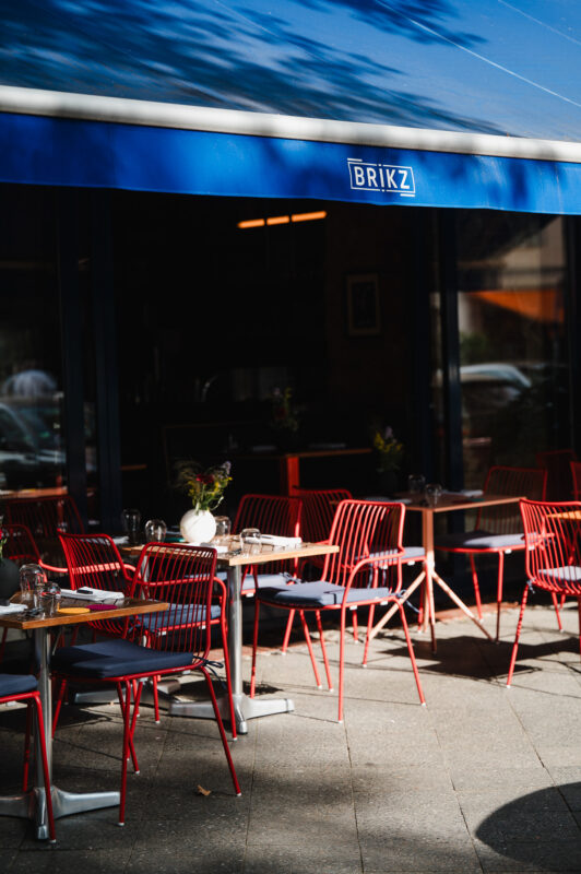 Red metal chairs and tables set up on the terrace of Restaurant Brikz, under a blue awning.