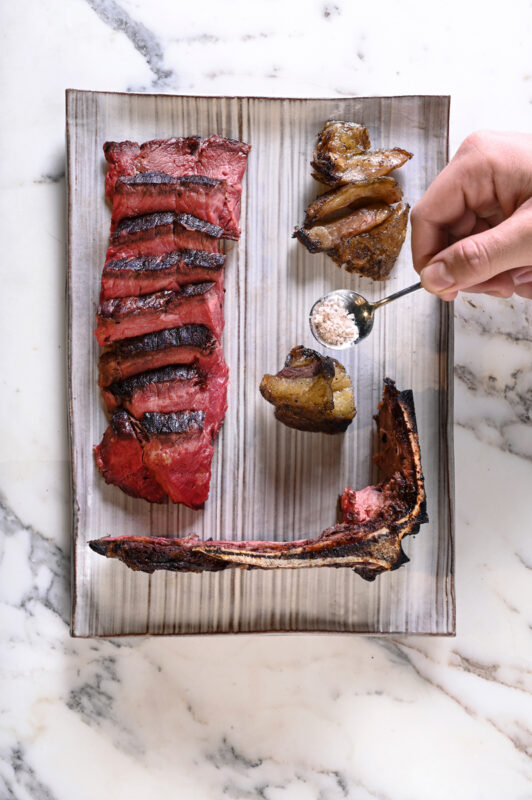 Grilled meat platter featuring sliced steak, roasted pieces, and a hand sprinkling salt on the dish.