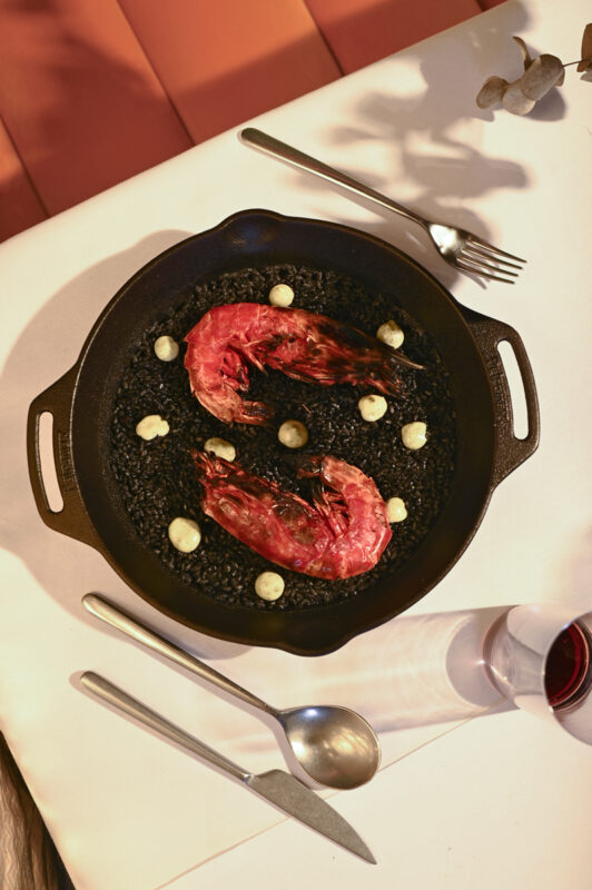 Black paella with two large grilled shrimp and dollops of sauce, set on a white tablecloth with cutlery.