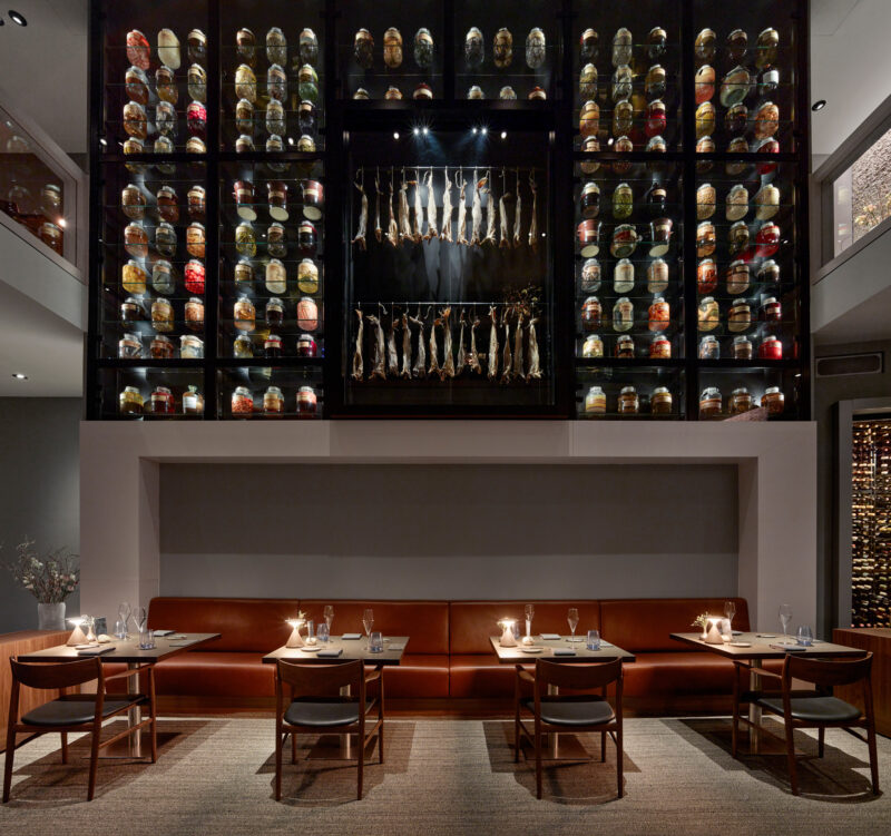 Modern restaurant interior featuring a wall of preserved jars and hanging fish, with wooden tables and chairs.