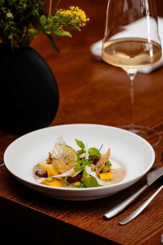 Zucchini and wild garlic dish garnished with cherry, herbs, and crispy elements, served with a glass of white wine.