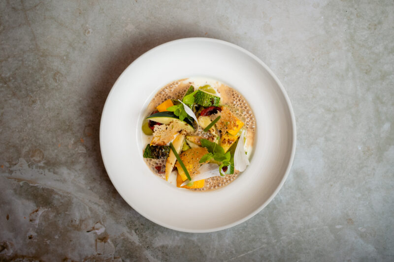 Colorful dish featuring zucchini, wild garlic, and cherries, garnished with greens, served in a white bowl on a textured s...