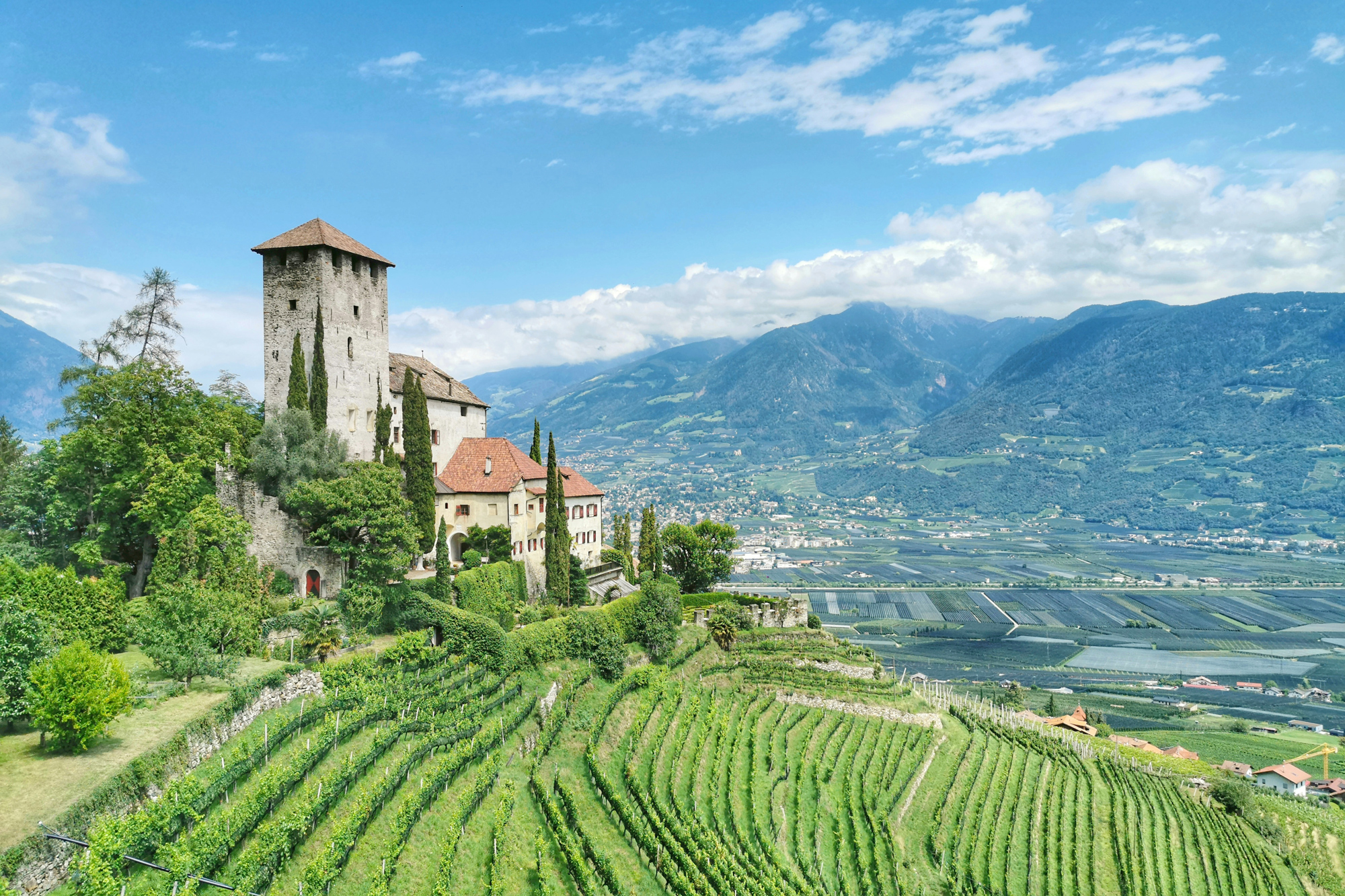 Castel Fragsburg with a stone tower, surrounded by lush greenery and vineyards, against a backdrop of mountains and blue sky.