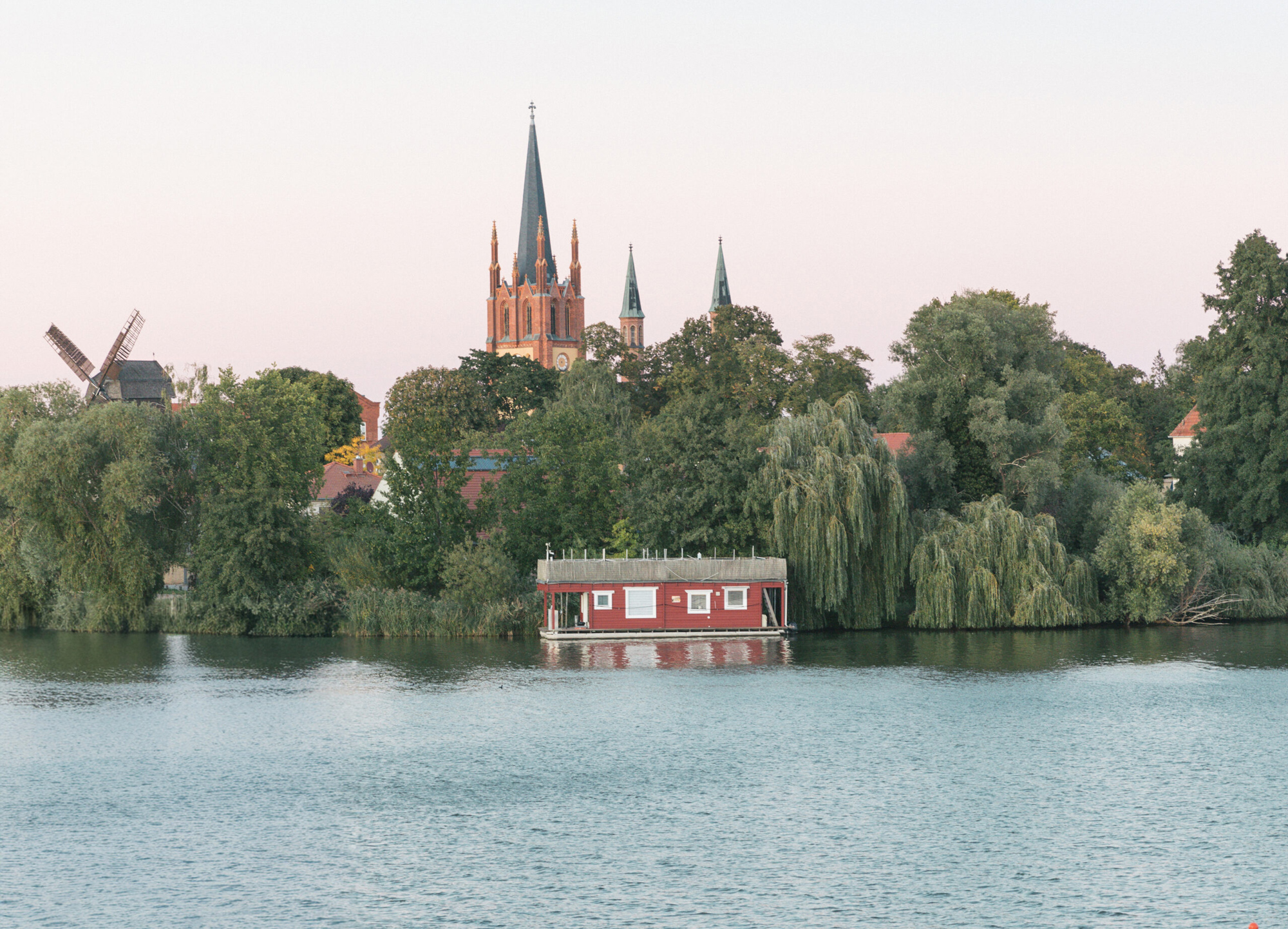 View of a red houseboat on a calm river, with a church spire and trees in the background, during sunset.