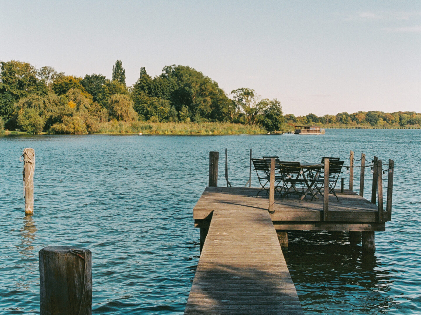 Wooden dock extending over water with a table and chairs, surrounded by trees and a clear blue sky.