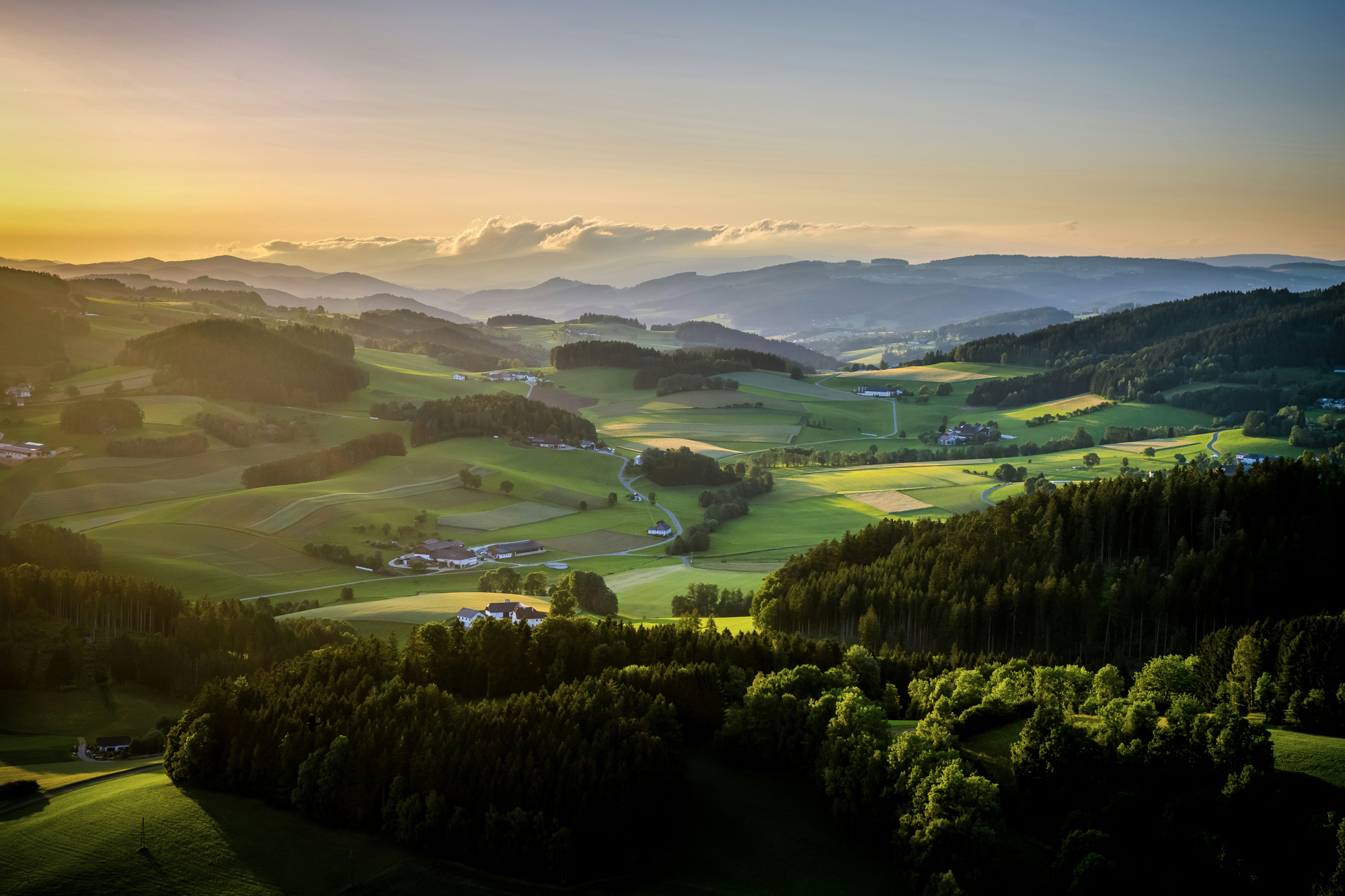 Lush green hills and valleys under a sunset sky, with scattered farms and forests in the foreground and background.