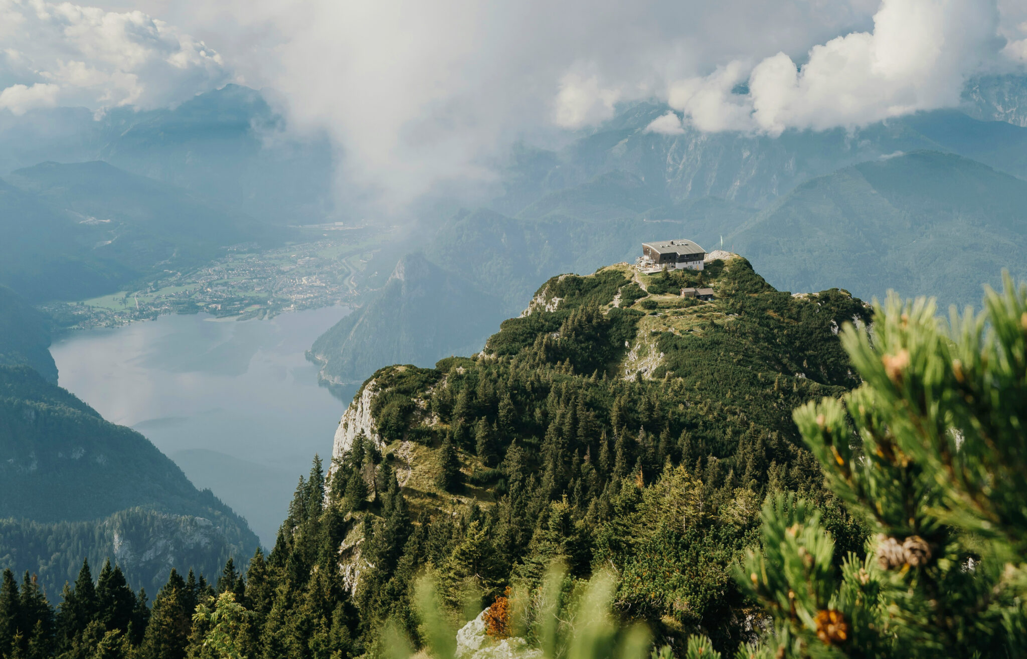 Mountain landscape featuring a house atop a rocky peak, overlooking a lake and valley, with clouds in the sky.