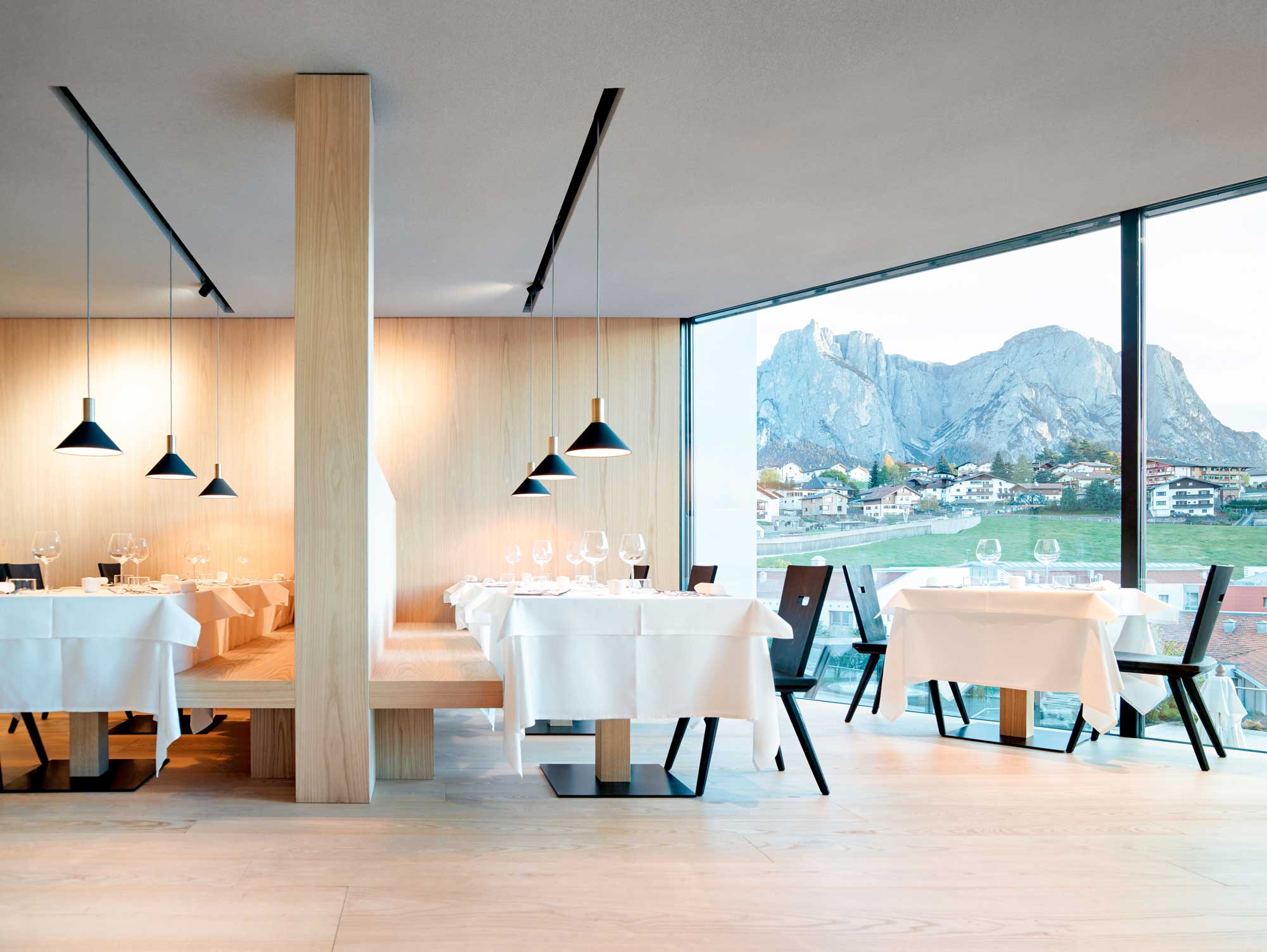 Modern restaurant interior with wooden accents, tables set with white linens, and a view of mountains through large windows.