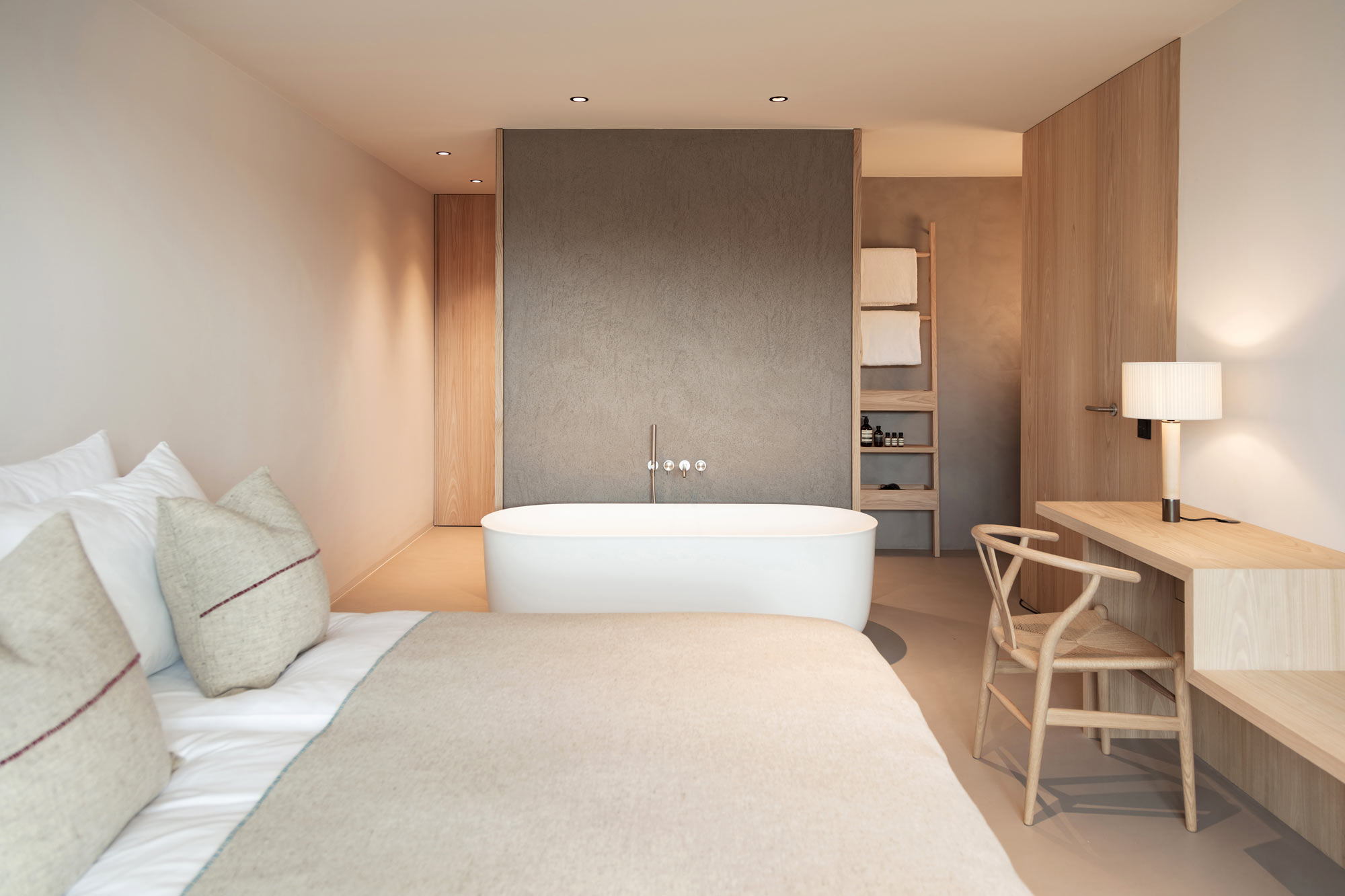 Modern hotel room featuring a bed with beige linens, a freestanding bathtub, a wooden desk, and minimalist decor.