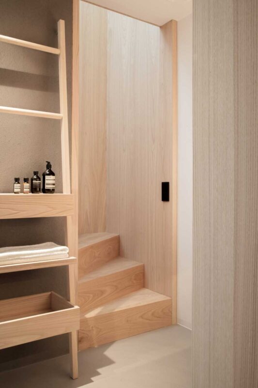 Wooden staircase leading to a light-filled area, with shelves displaying toiletries and neatly folded towels.
