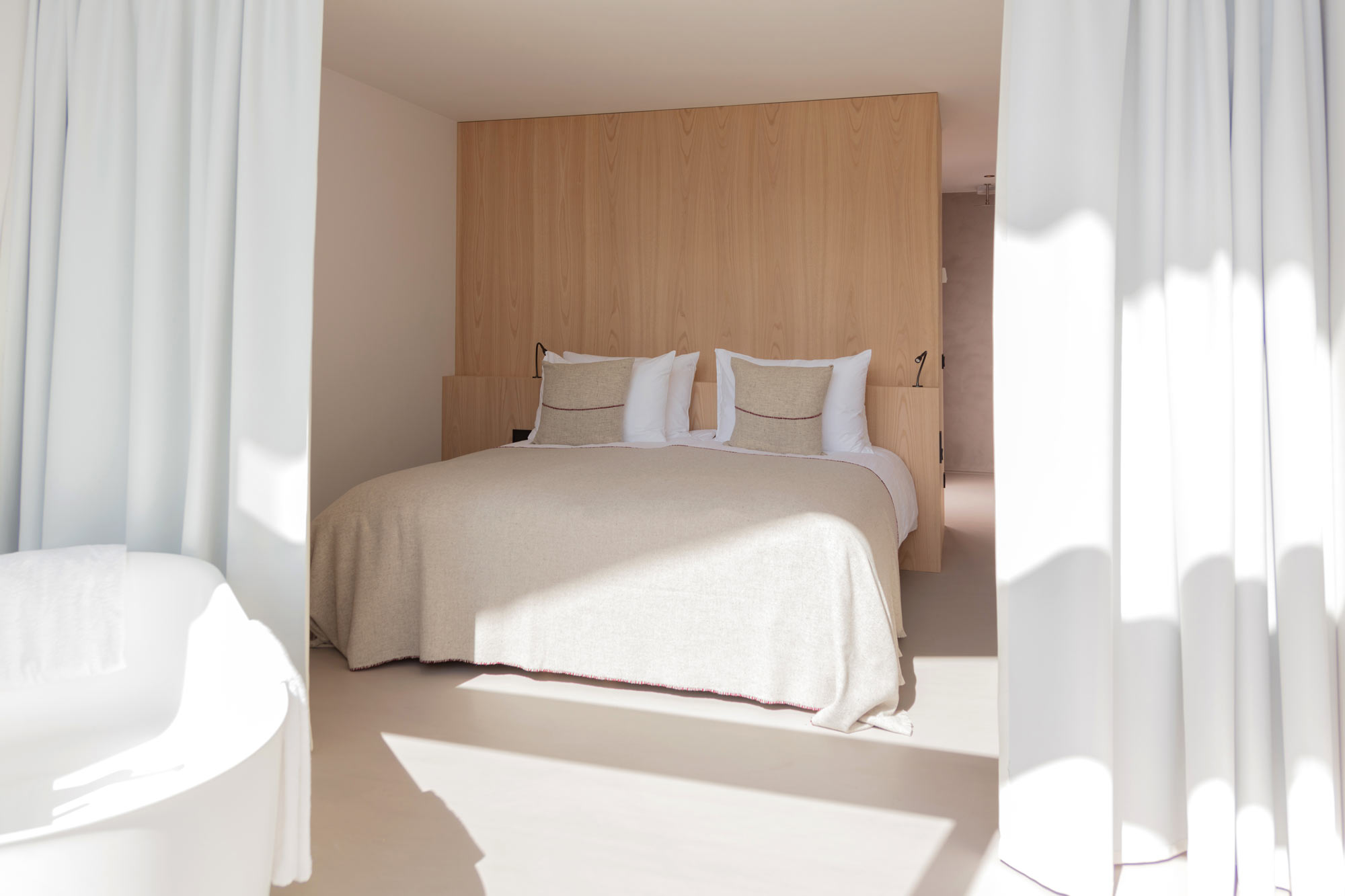 Modern hotel room featuring a large bed with neutral-toned linens, wooden paneling, and bright natural light.