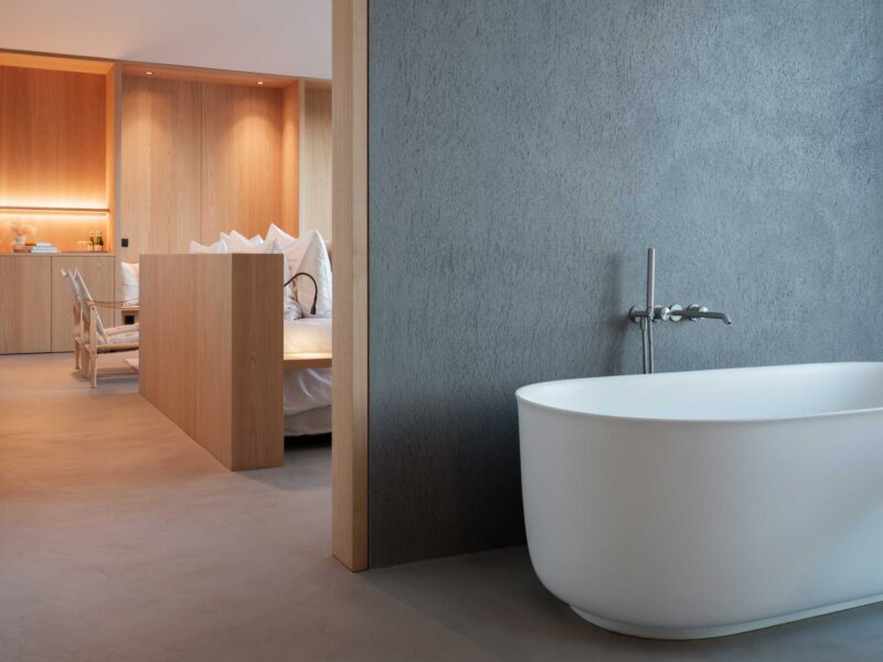 Modern hotel room featuring a freestanding white bathtub in the foreground, with wooden accents and a bed visible in the b...