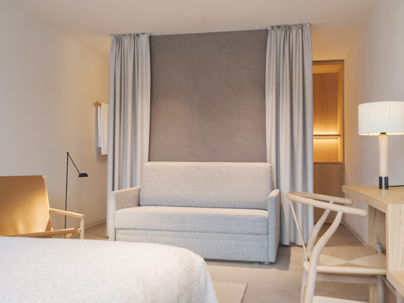 Modern hotel room featuring a gray sofa, wooden desk, and soft lighting, with neutral tones and elegant decor.